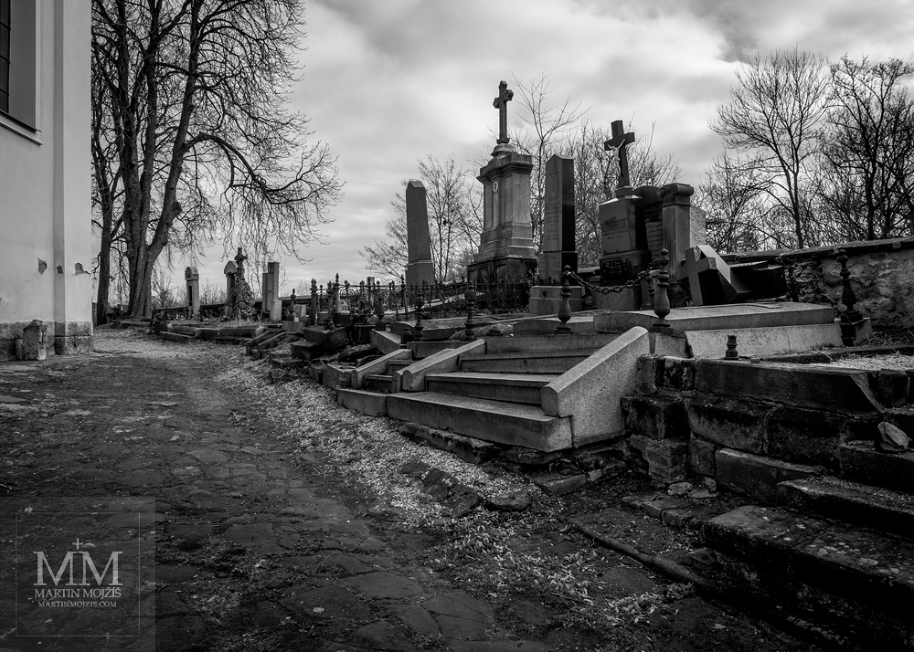 Hřbitov u kostela, černobílá fotografie. Fotografie vytvořena objektivem Olympus 12 - 40 mm 2.8 Pro.
